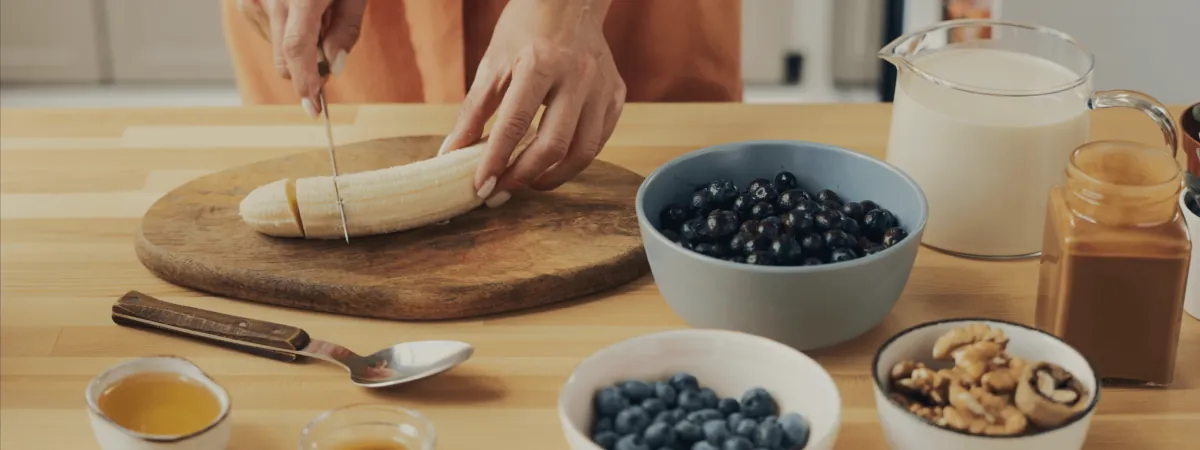 Hands slicing a banana beside bowls of blueberries and walnuts