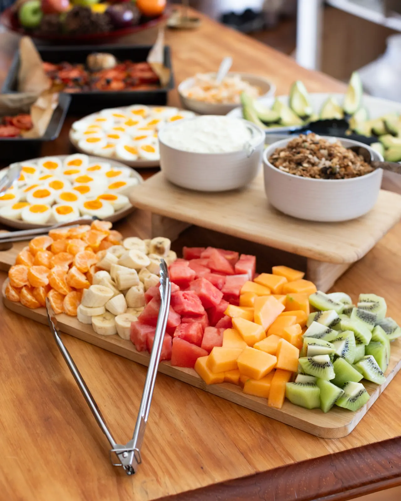 A breakfast board of melon, kiwi fruit, banana and orange, with yoghurt, granola and boiled eggs behind