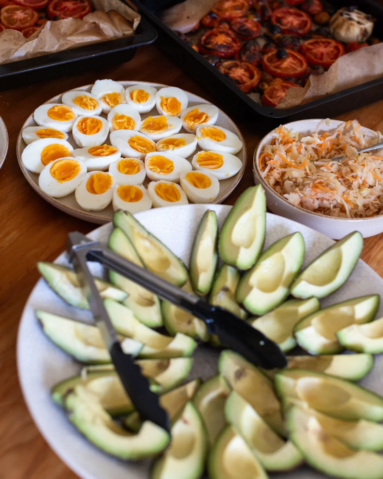 A plate of sliced avocado, with boiled eggs, trays of roast tomatoes and a bowl of slaw behind