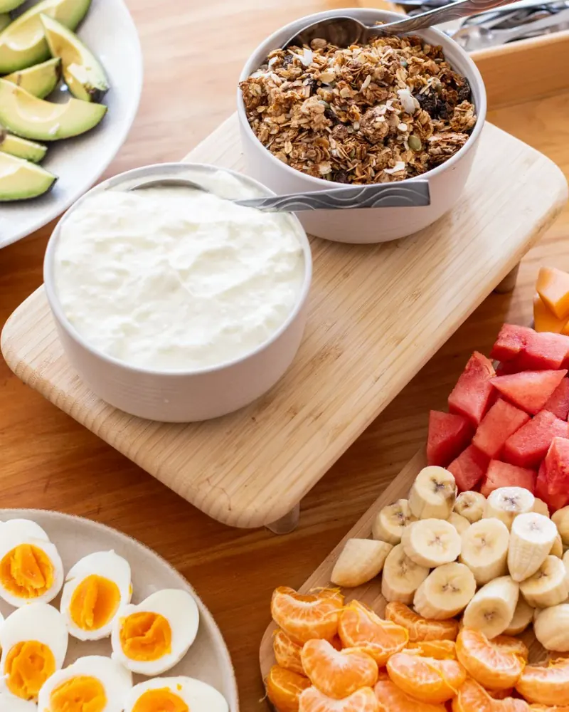 Bowls of yoghurt and granola beside sliced avocado, boiled eggs and cut fruit