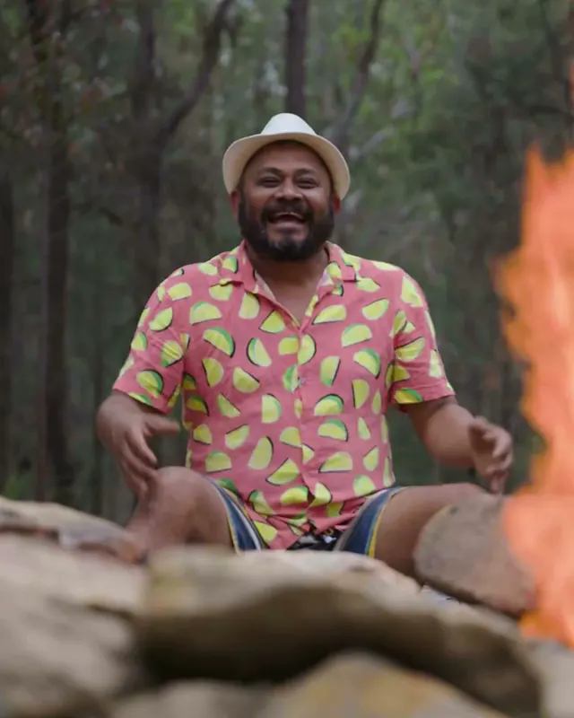 Jeevi, a guest, laughing on the rocks by the fire pit in a straw hat and a pink shirt printed with limes