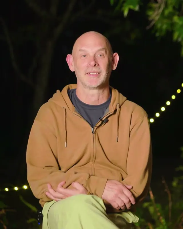 Mark, a guest, smiling, sitting outdoors at dusk in a tan hoodie with string lights behind him